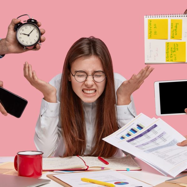 Photo of depressed female has deadline and much work, spreads hands, has long hair clenches teeth with dissatisfaction, poses at desktop with papers, models against pink background. Negative emotions
