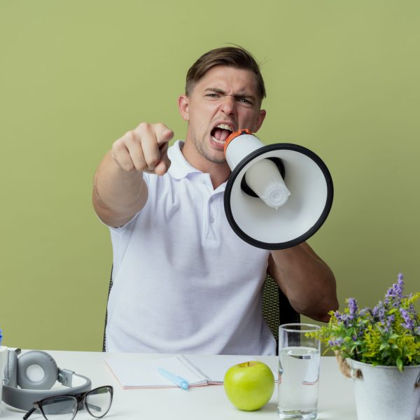 confident young handsome male student sitting at desk with school tools speaks on loudspeaker and showing you gesture isolated on olive green