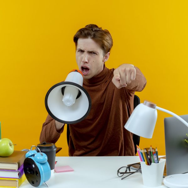 confident young student boy sitting at desk with school tools speaks on loudspeaker and showing you gesture isolated on yellow background