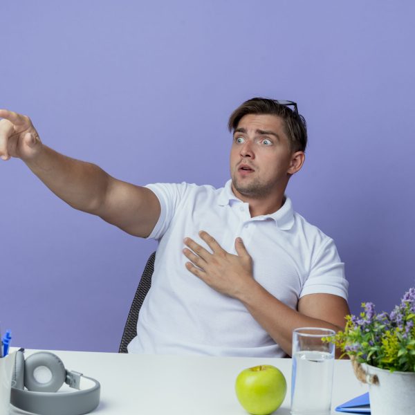 looking at side scared young handsome male student sitting at desk with school tools points at side isolated on blue background