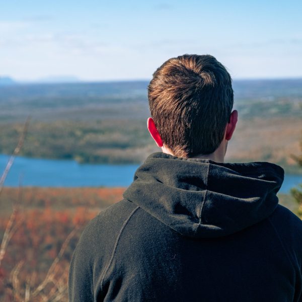 A man with a hooded jacket resting on a hill looking at  a beautiful river landscape on a sunny day