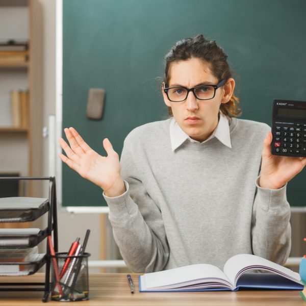 unpleased spreading hands young male teacher holding calculator sitting at desk with school tools on in classroom